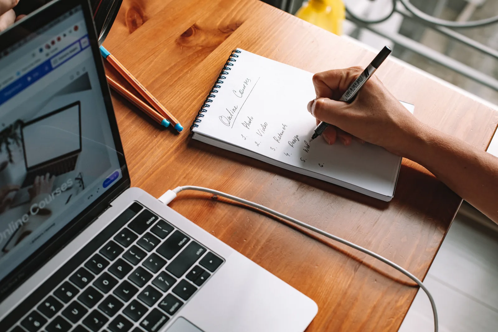 Person writing notes on a notepad beside an open laptop