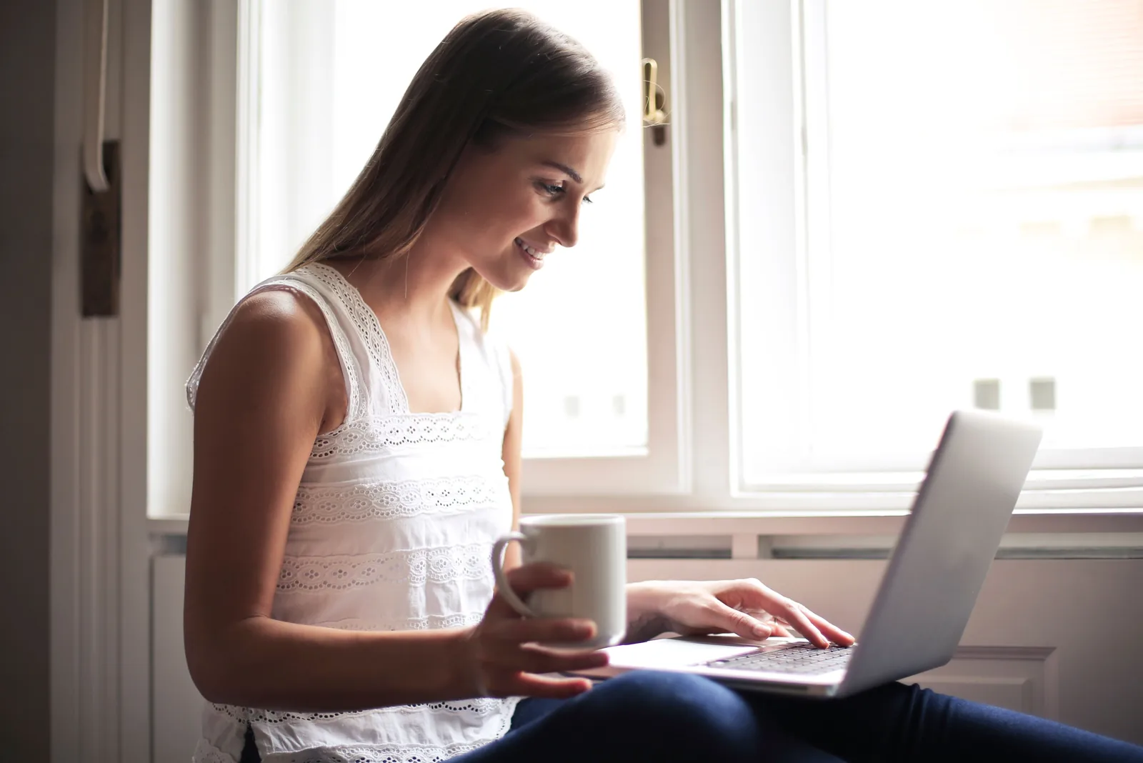 Woman smiling at her laptop by a bright window, holding a mug