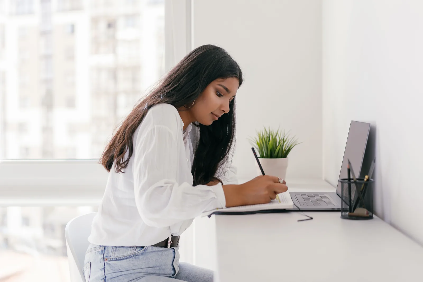 Woman writing in a notebook in front of her laptop at a home desk